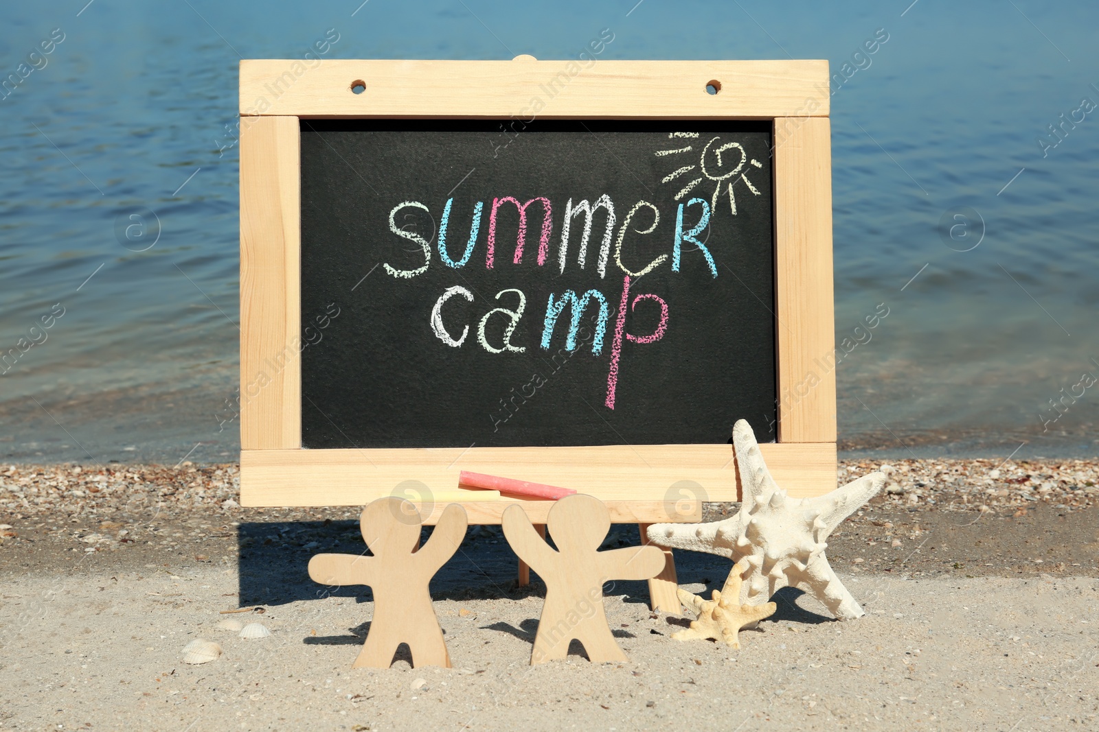 Photo of Small chalkboard with text SUMMER CAMP, wooden people and starfish on beach near river