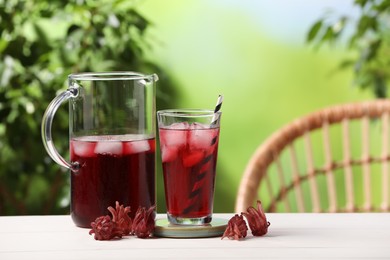 Photo of Refreshing hibiscus tea with ice cubes and roselle flowers on white wooden table against blurred green background. Space for text