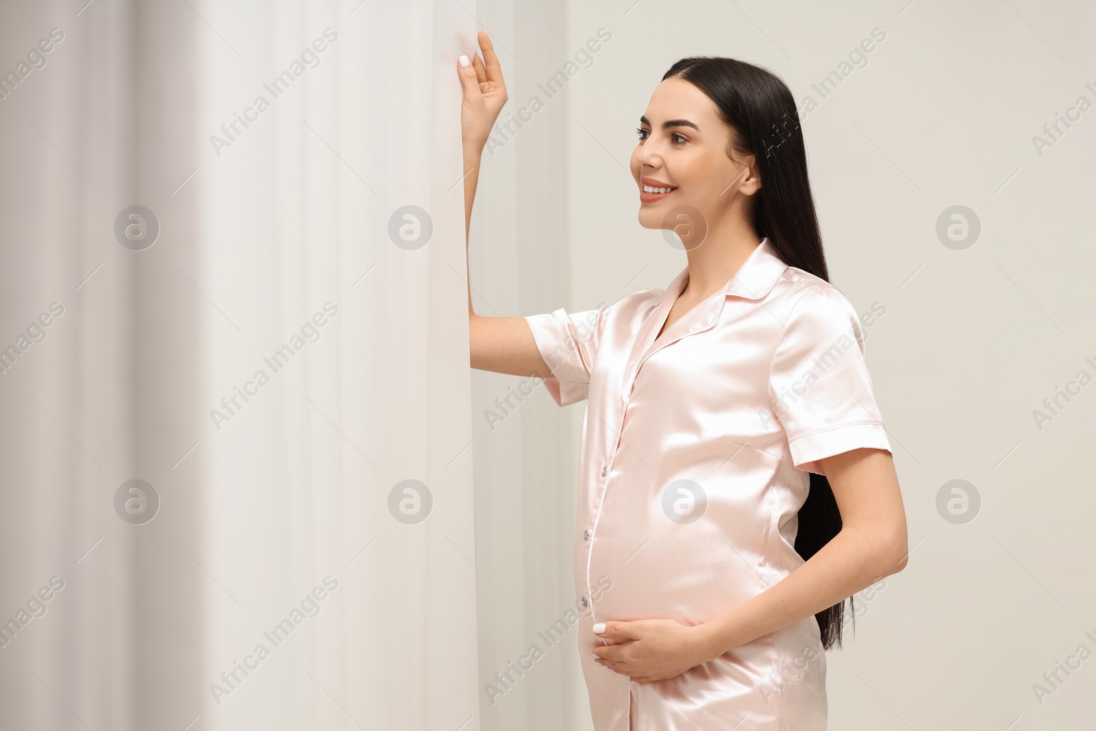 Photo of Happy pregnant woman in pajamas near window indoors