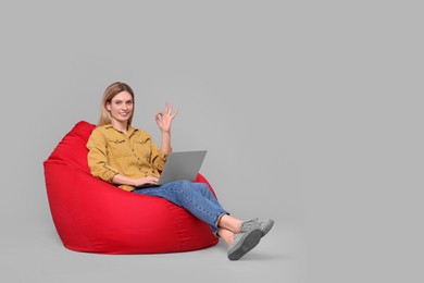 Photo of Happy woman with laptop sitting on beanbag chair and showing ok gesture against light grey background. Space for text