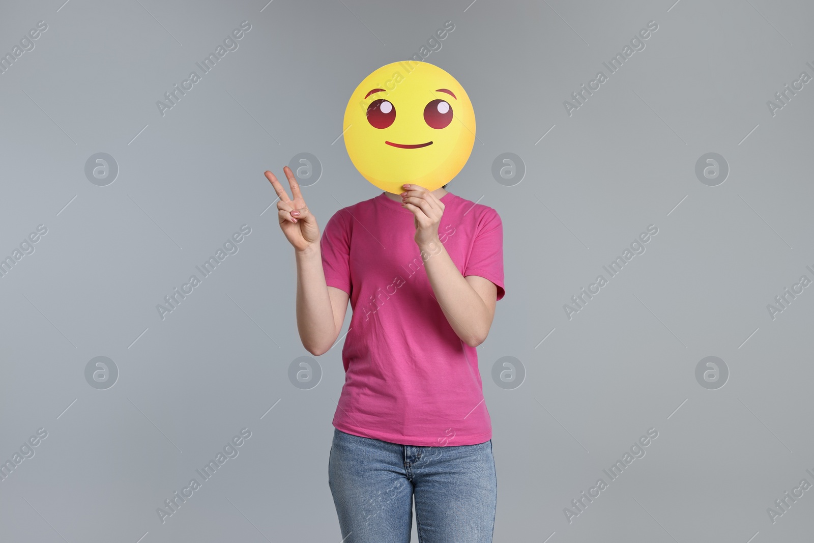 Photo of Woman covering face with smiling emoticon and showing peace sign on grey background