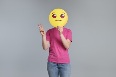 Woman covering face with smiling emoticon and showing peace sign on grey background