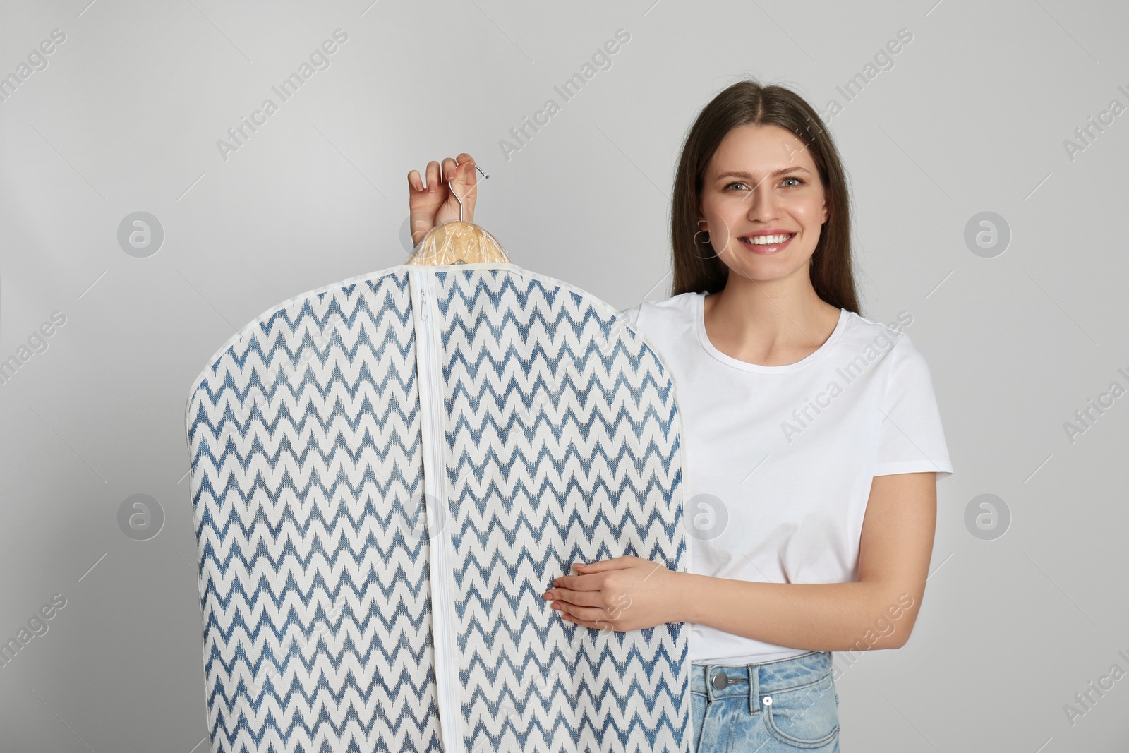 Photo of Young woman holding hanger with clothes in garment cover on light grey background. Dry-cleaning service