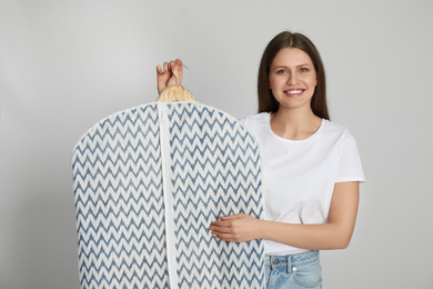 Young woman holding hanger with clothes in garment cover on light grey background. Dry-cleaning service