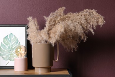 Photo of Ceramic vase with fluffy dry plants, painting and modern night lamp on wooden table near brown wall