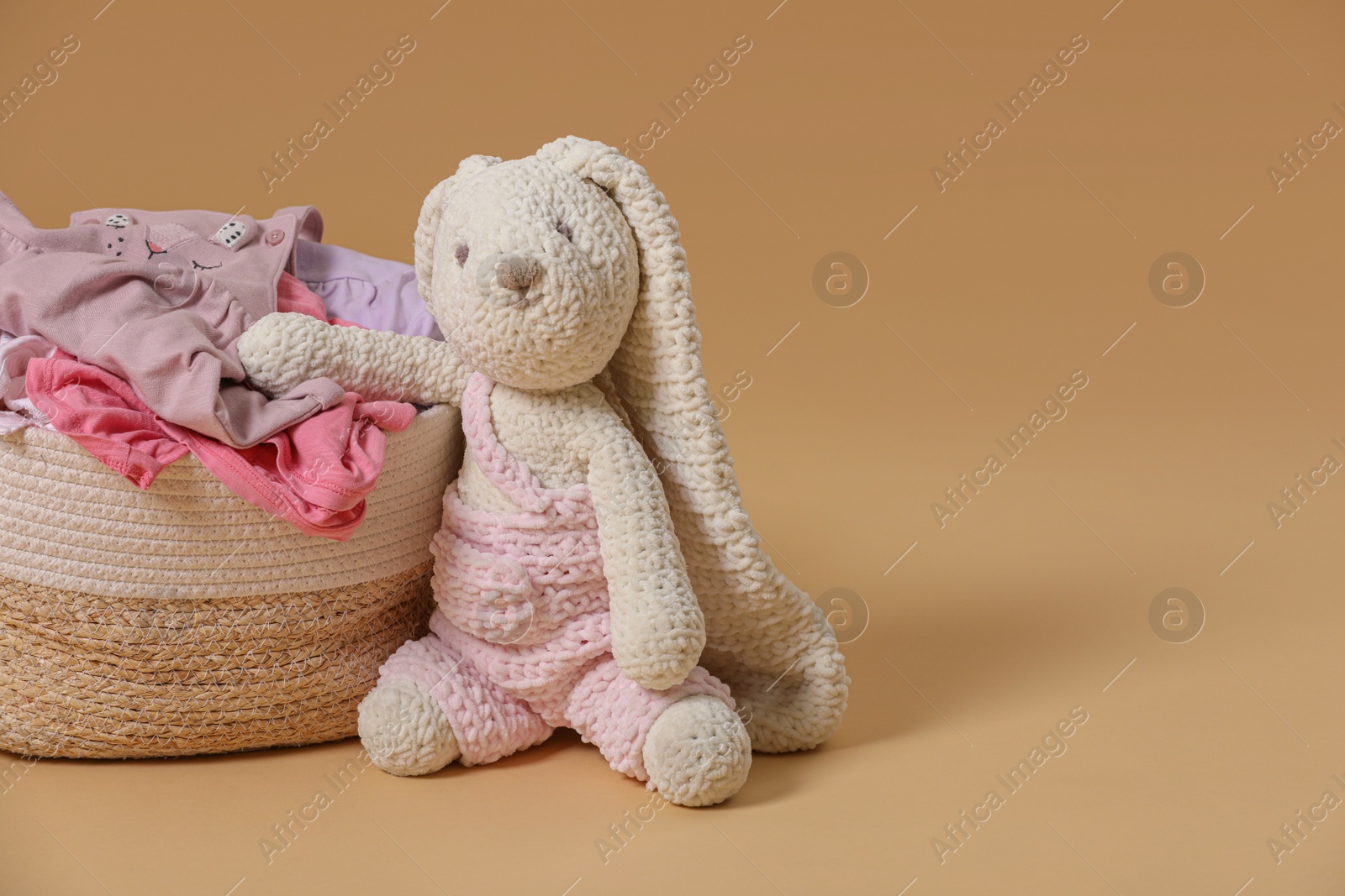 Photo of Laundry basket with baby clothes near soft toy on light brown background. Space for text