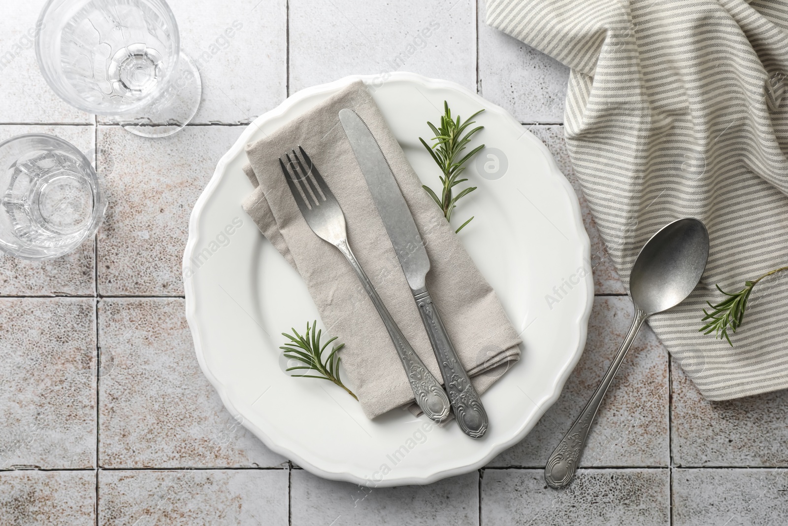 Photo of Stylish setting with cutlery, napkin, rosemary and plate on light tiled table, top view