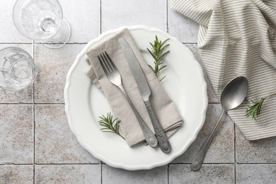 Stylish setting with cutlery, napkin, rosemary and plate on light tiled table, top view