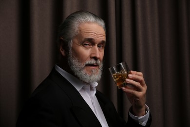 Senior man in formal suit holding glass of whiskey with ice cubes on brown background