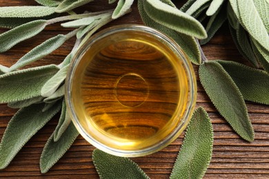 Bowl of essential sage oil and leaves on wooden table, flat lay