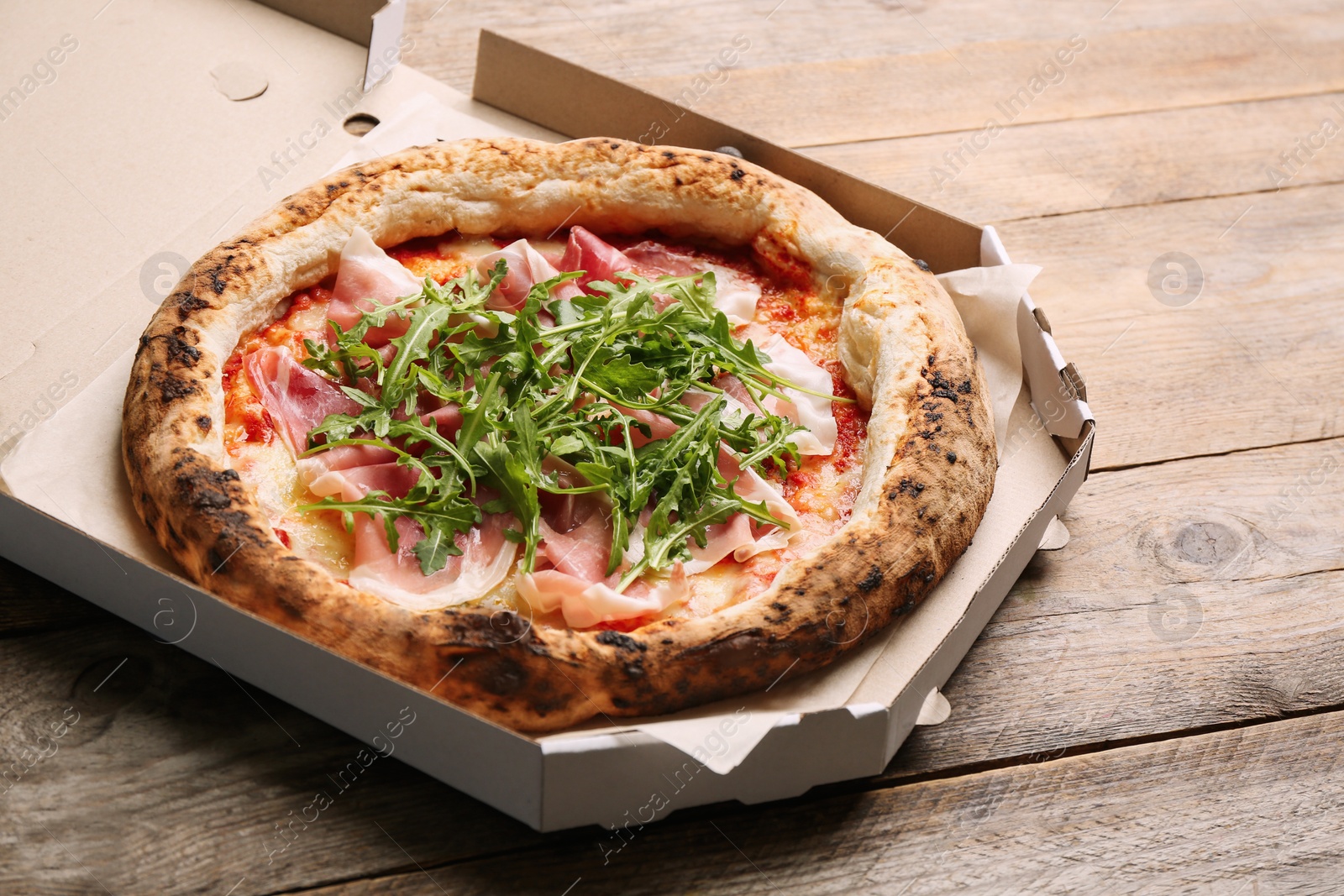 Photo of Tasty pizza with meat and arugula in cardboard box on wooden table