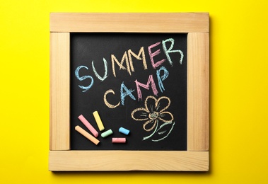 Small blackboard with text SUMMER CAMP, drawing and chalk sticks on color background, top view