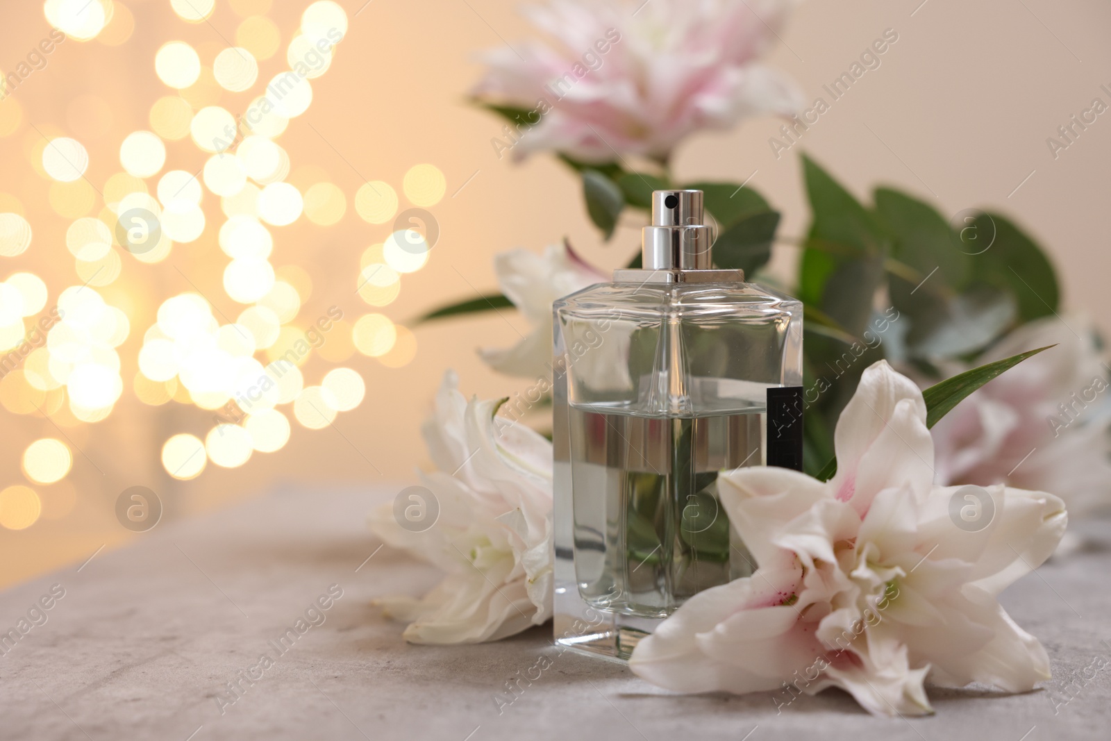 Photo of Bottle of perfume and beautiful lily flowers on table against beige background with blurred lights, closeup. Space for text