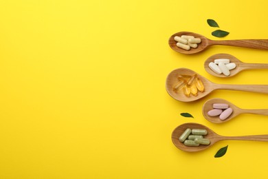 Different vitamin pills in spoons and green leaves on yellow background, flat lay. Space for text