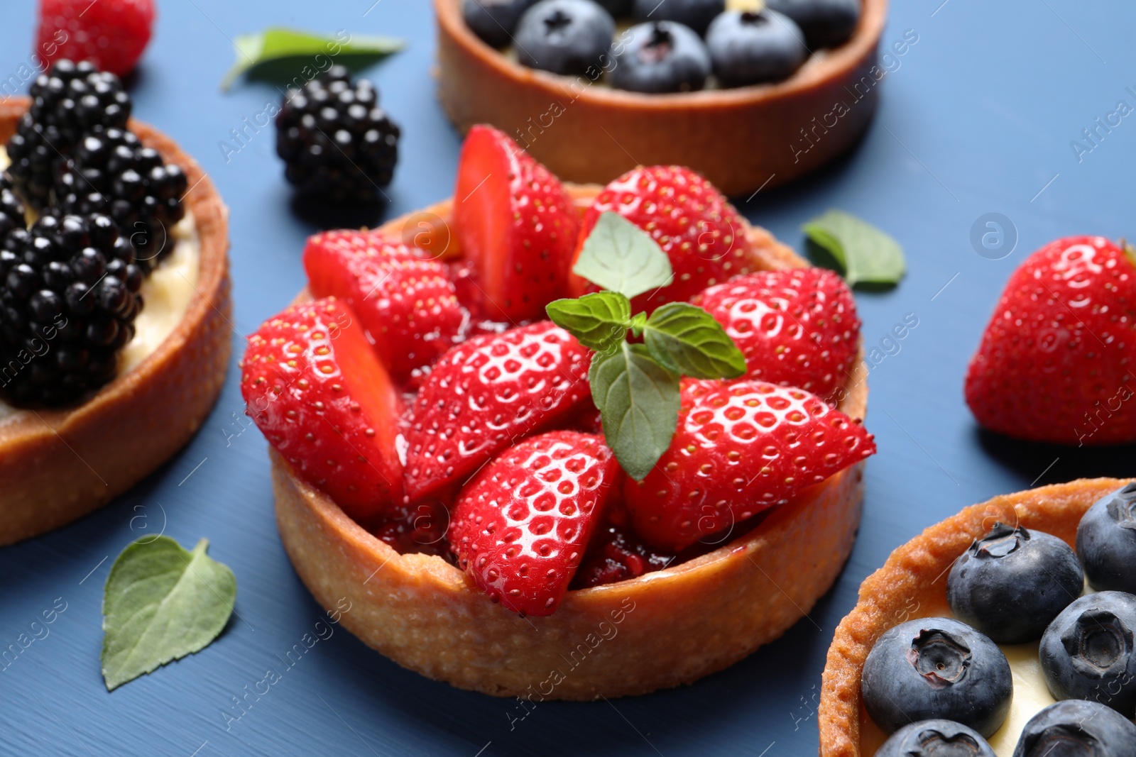 Photo of Tartlets with different fresh berries and mint on blue wooden table, closeup. Delicious dessert