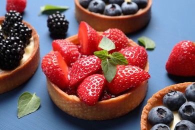 Photo of Tartlets with different fresh berries and mint on blue wooden table, closeup. Delicious dessert