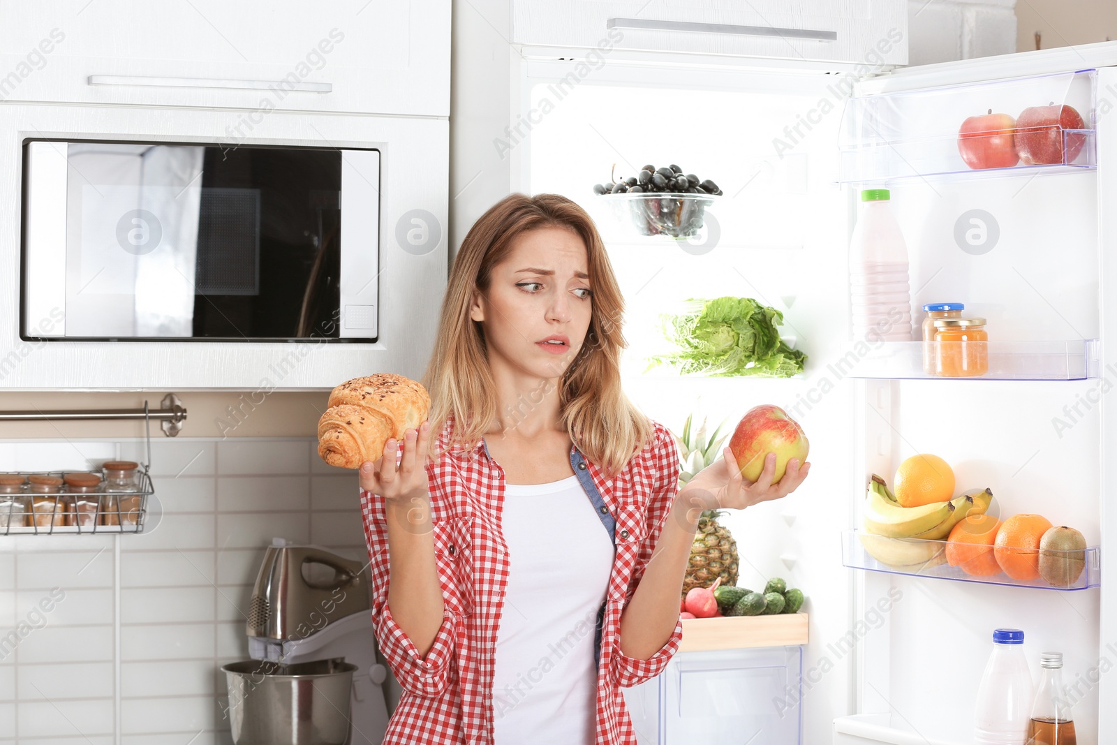 Photo of Woman choosing between apple and croissant near fridge at kitchen. Healthy diet
