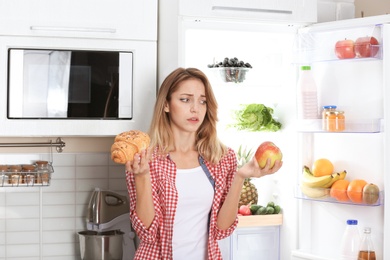 Woman choosing between apple and croissant near fridge at kitchen. Healthy diet