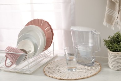 Photo of Drainer with different clean dishware, cup, glass and filter jug on light table indoors