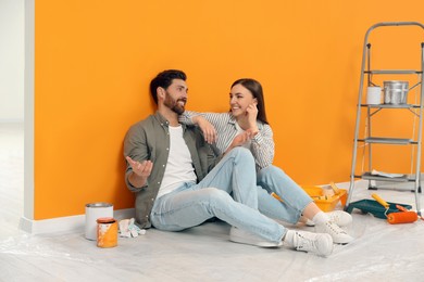 Photo of Happy designers sitting on floor with painting equipment near freshly painted orange wall indoors