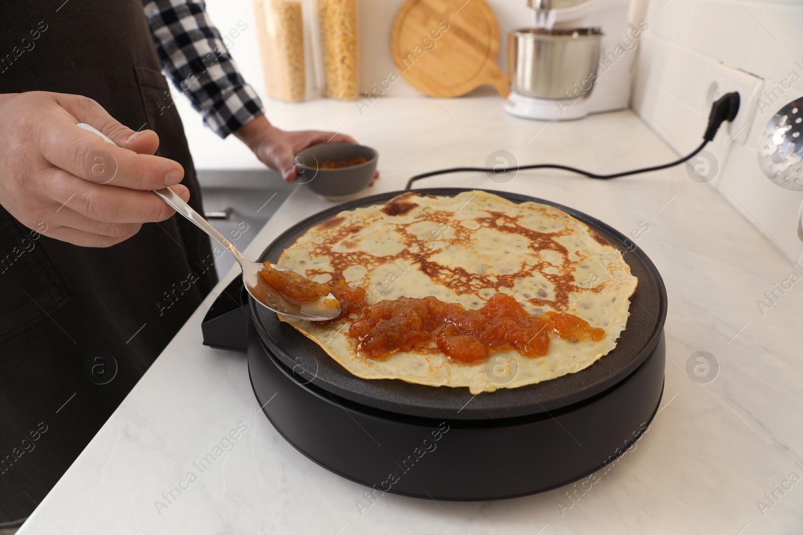 Photo of Man cooking delicious crepe with jam on electric pancake maker in kitchen, closeup