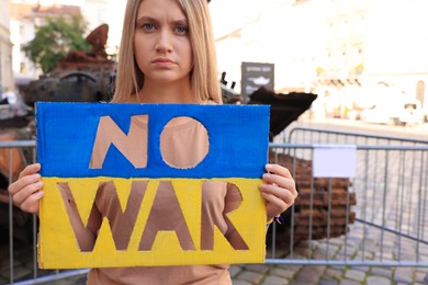 Photo of Sad woman holding poster in colors of Ukrainian flag with words No War near broken tank on city street