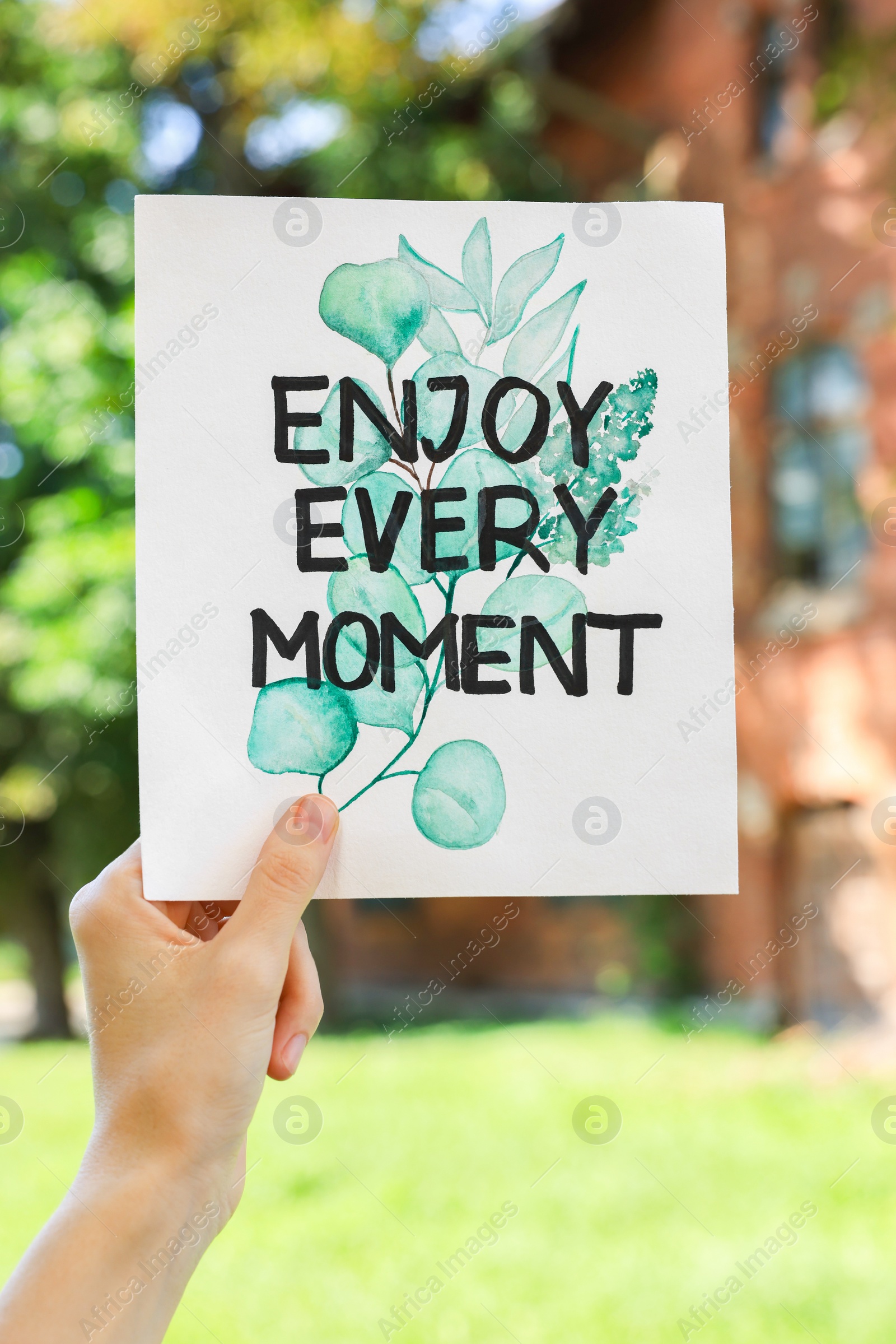 Photo of Woman holding card with phrase Enjoy Every Moment outdoors, closeup