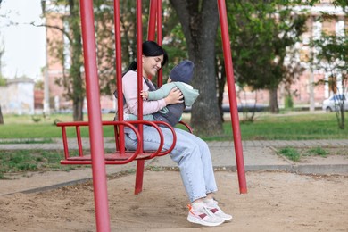 Mother holding her child in sling (baby carrier) on swing in park