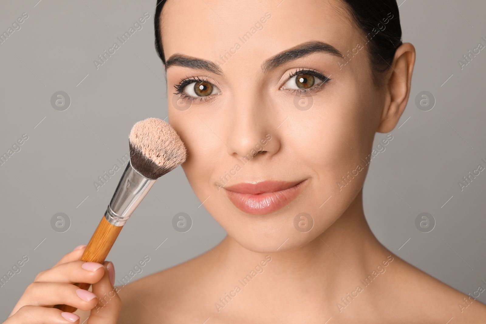 Photo of Beautiful young woman applying face powder with brush on grey background, closeup