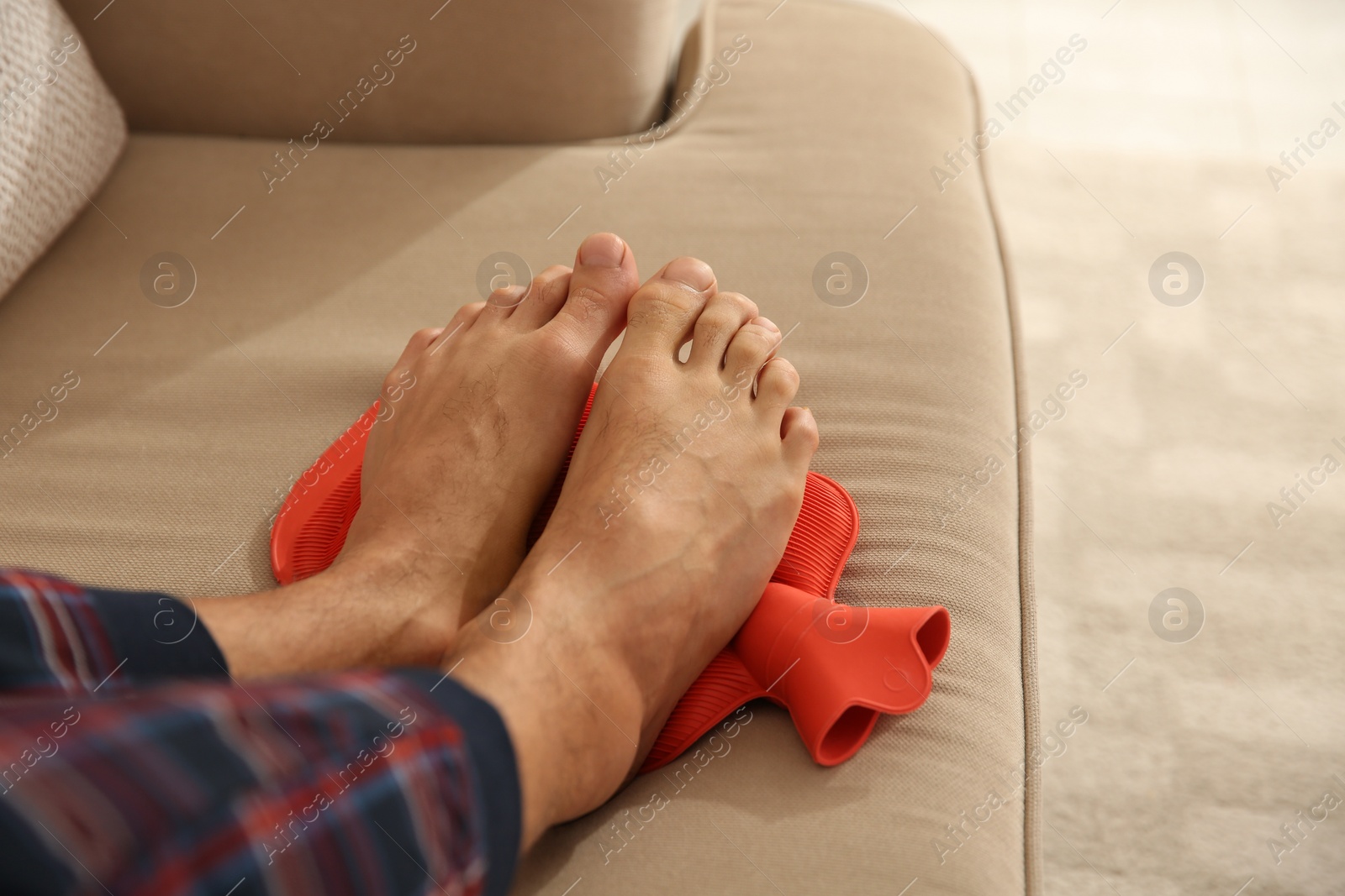 Photo of Man warming feet with hot water bottle on sofa, closeup