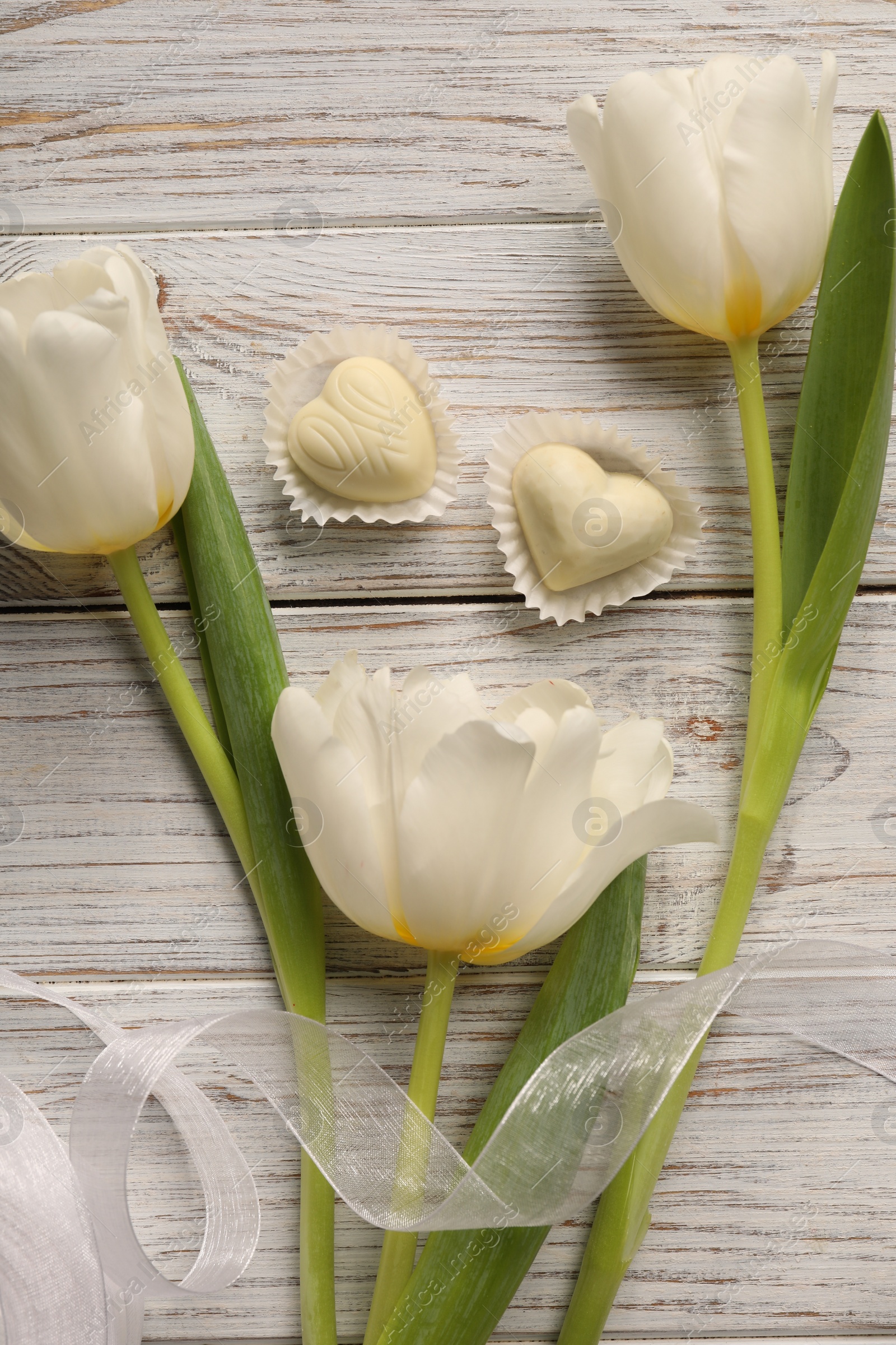 Photo of Delicious heart shaped chocolate candies and beautiful tulips on white wooden table, flat lay