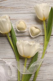 Delicious heart shaped chocolate candies and beautiful tulips on white wooden table, flat lay