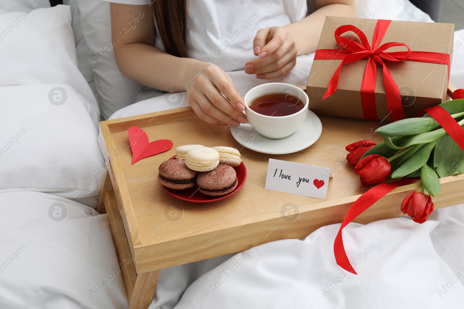 Photo of Tasty breakfast served in bed. Woman with tea, macarons, gift box, flowers and I Love You card at home, closeup