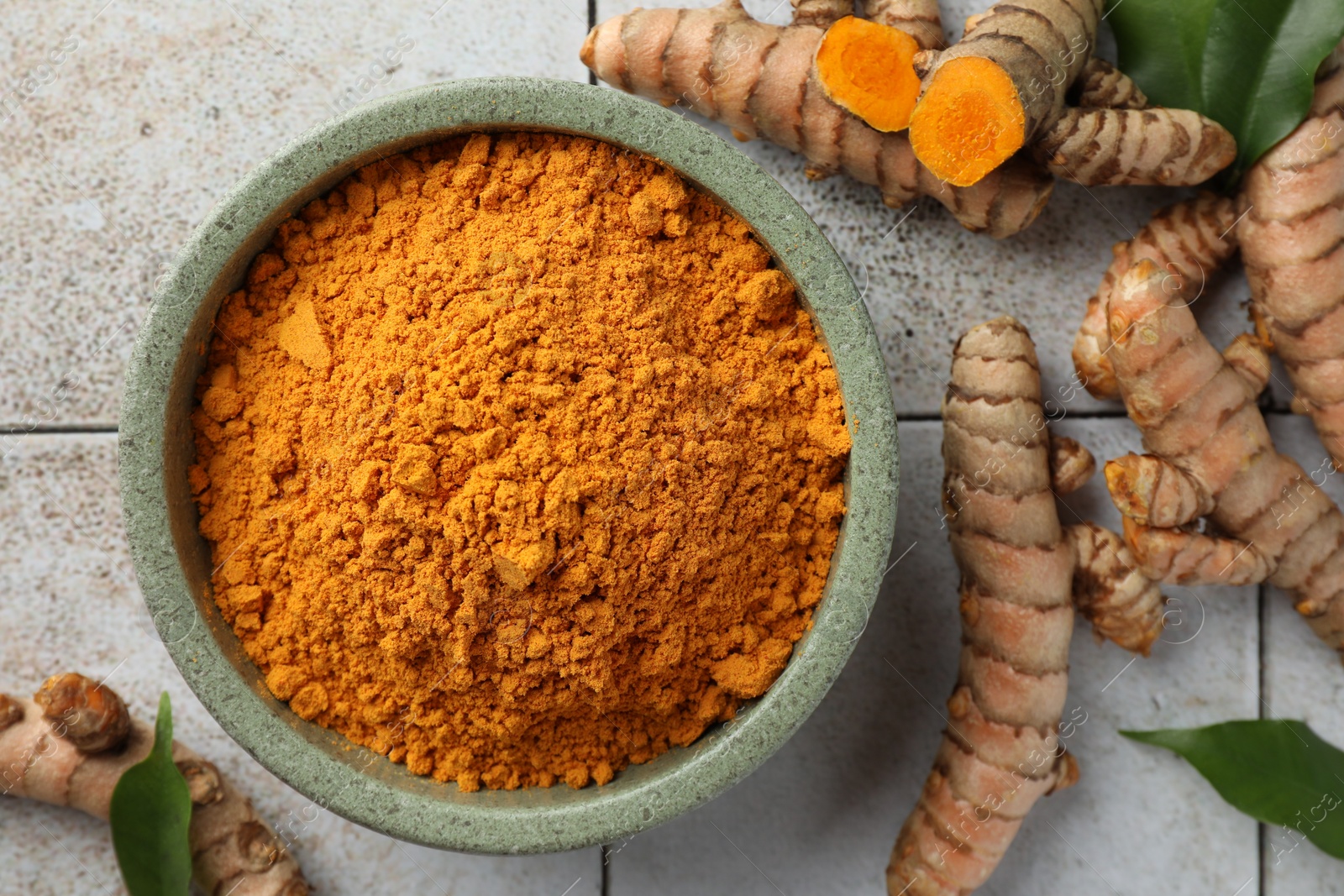 Photo of Aromatic turmeric powder, raw roots and green leaves on light tiled table, flat lay