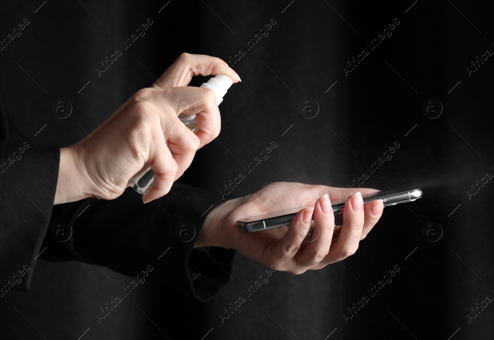 Photo of Woman spraying antiseptic onto smartphone against black background, closeup