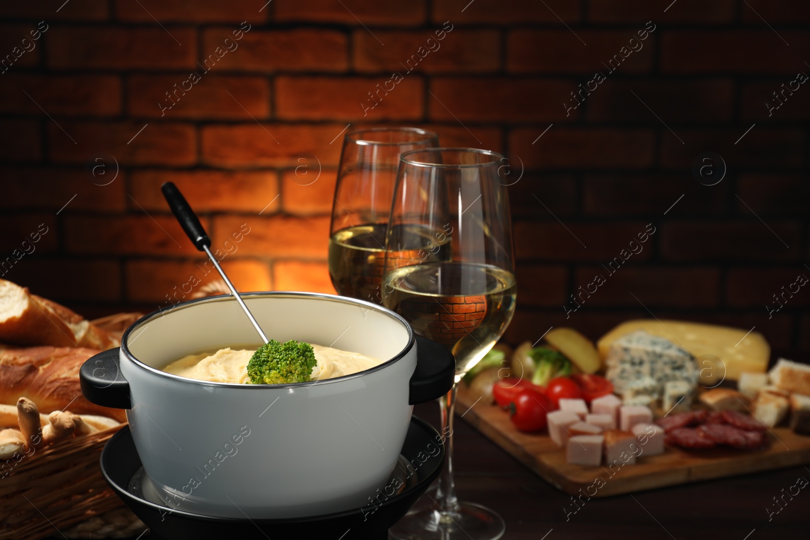 Photo of Fork with piece of broccoli, melted cheese in fondue pot, wine and snacks on table, closeup