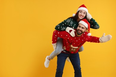 Young couple in Christmas sweaters, Santa hats and knitted mittens on orange background. Space for text
