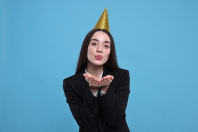 Happy woman in party hat blowing kiss on light blue background