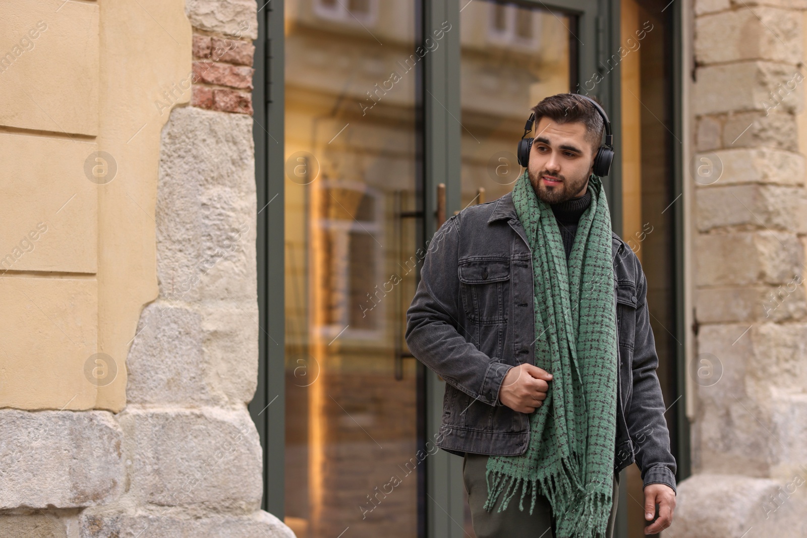 Photo of Handsome man in warm scarf and headphones near building outdoors. Space for text