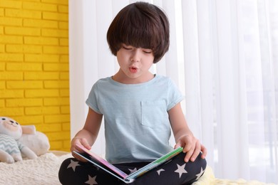 Photo of Cute little boy reading book on bed at home