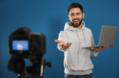 Photo of Young blogger with laptop recording video on camera against blue background