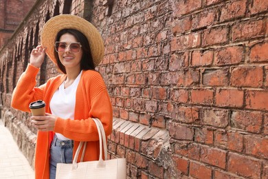 Photo of Young woman with stylish bag and cup of hot drink near red brick wall outdoors, space for text