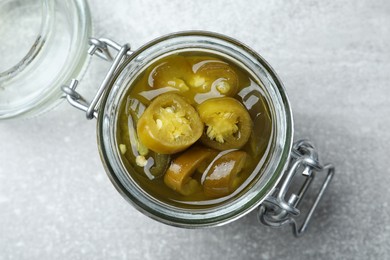 Glass jar with slices of pickled green jalapeno peppers on light grey table, top view