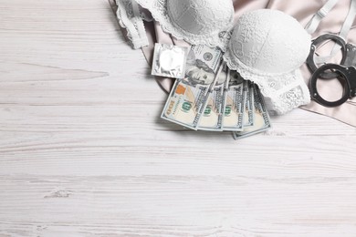 Photo of Prostitution concept. Handcuffs, dollar banknotes, condom and bra on white wooden table, flat lay. Space for text