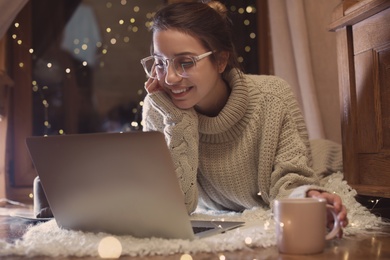 Woman with cup of hot beverage using laptop at home in winter evening