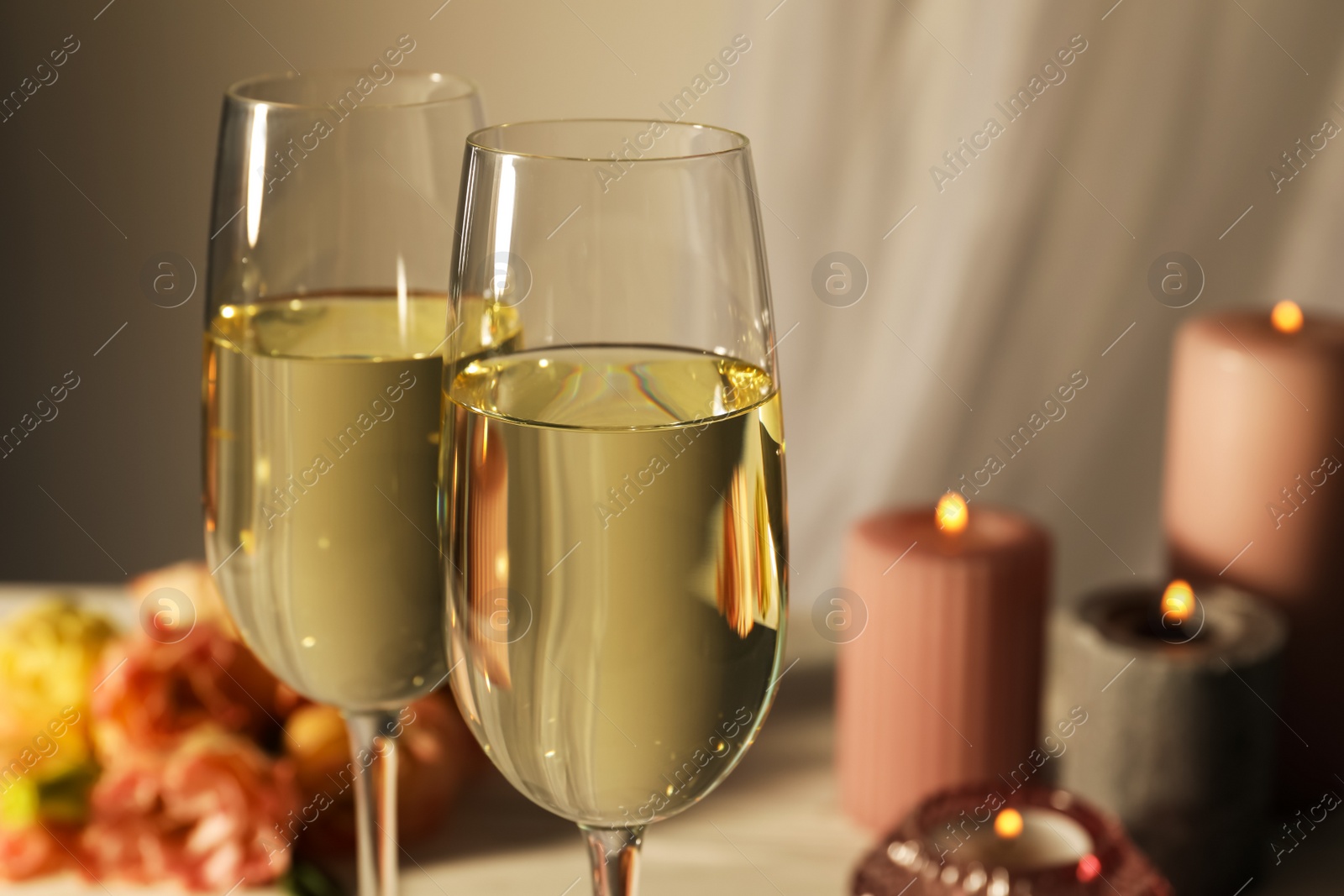 Photo of Glasses of sparkling wine, flowers and burning candles on white table, closeup