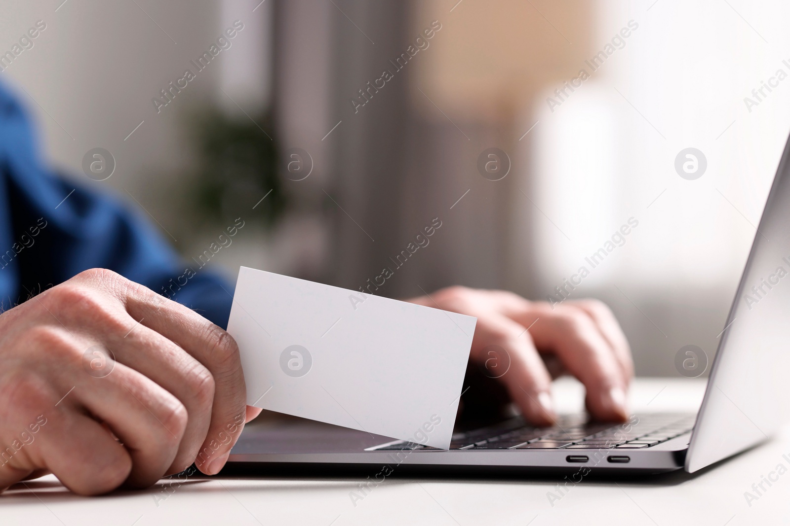 Photo of Man with laptop holding blank business card at white table indoors, closeup