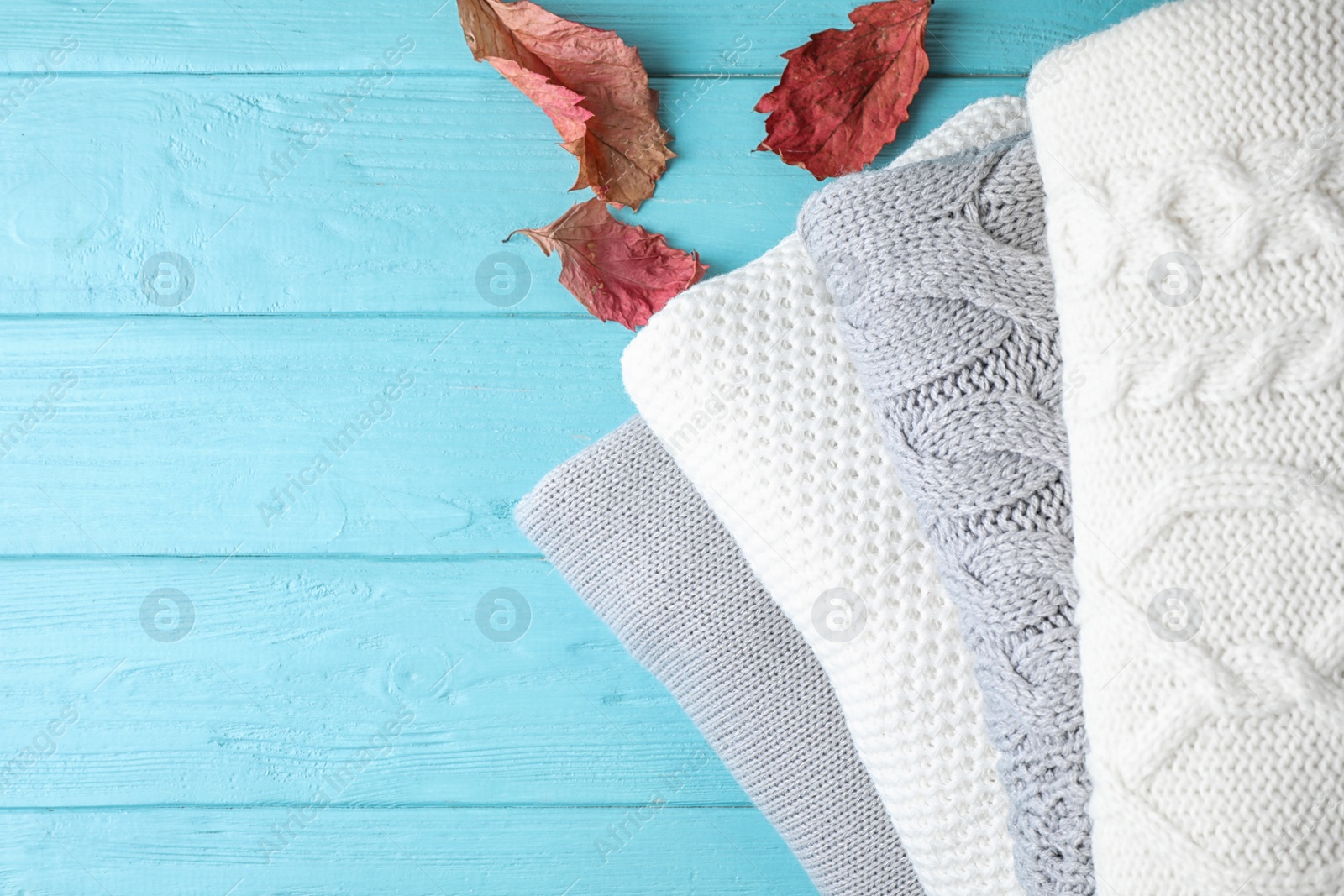 Image of Stack of folded warm sweaters and dry leaves on turquoise wooden background, top view. Space for text