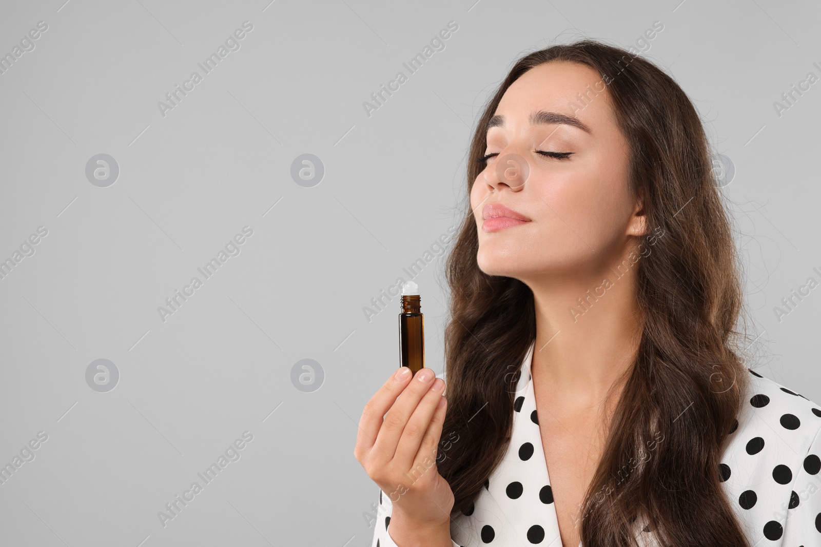 Photo of Beautiful young woman with roller bottle of essential oil on light grey background, space for text