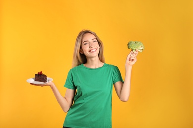 Woman choosing between cake and healthy broccoli on yellow background